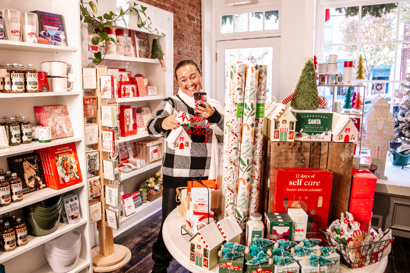 In Duly Noted shop all decked out for the holidays, a person stands behind a rotating display table covered with Christmas gifts and decorations, smiling while holding a small item, with shelves of wrapped products in the background.
