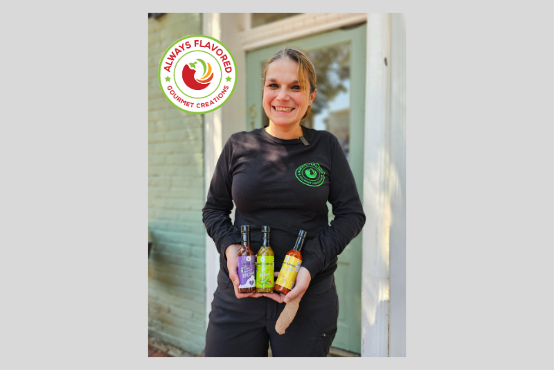 Smiling person in a black long-sleeve shirt holding three bottles with purple, green, and yellow labels, standing in front of a glass door and light brick wall.