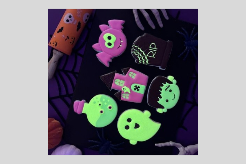 Colorful Halloween-themed cartoon ghosts and monsters in neon green and pink, with dark purple bats and spider webs, scattered around a purple background.