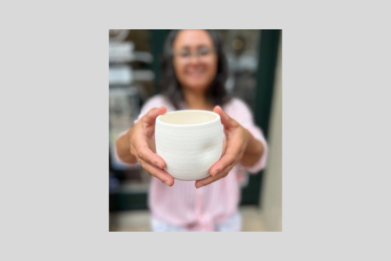 A person holds a white ceramic cup with both hands toward the camera, standing in a softly blurred indoor background with shelves and a green door frame.