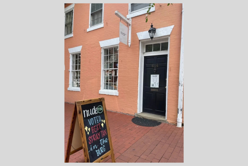 Brick building entrance with a dark door marked “614,” a wall-mounted lantern, and a sidewalk sign advertising “NudeFx” spray tan in burg and best spray tan in the Burg.